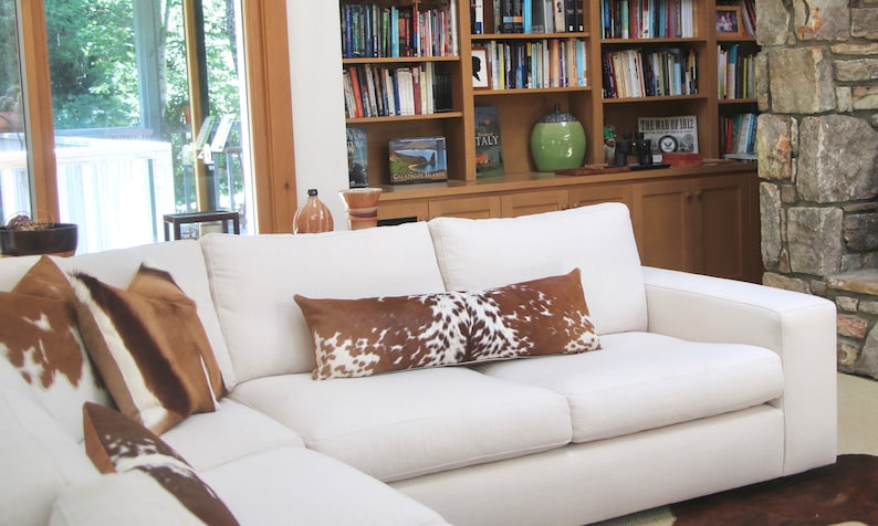 May include: A white sectional sofa with a brown and white cowhide patterned throw pillow and two square pillows. The sofa is in a living room with a bookshelf and a stone fireplace in the background.