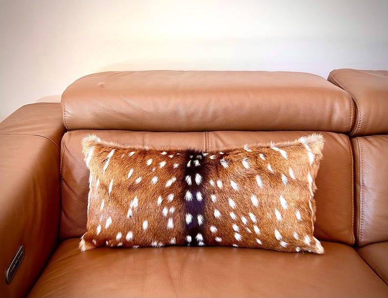 May include: A rectangular decorative pillow with a deer hide pattern. The pillow features a brown and white spotted design with a dark brown stripe down the center. The pillow is placed on a brown leather sofa.
