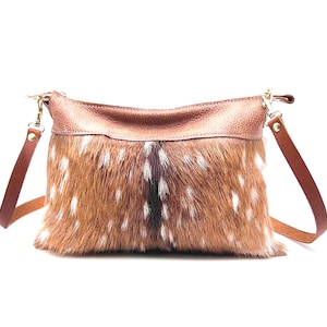 May include: A brown and white spotted crossbody bag with a leather top and straps. The bag has a zipper closure and gold-colored hardware. The bag's body is made of a spotted fur-like material, and the straps are a dark brown.