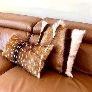May include: Two decorative throw pillows with natural fur covers. One pillow features a brown and white spotted pattern, while the other has a brown, white, and black striped design. The pillows rest on a brown leather sofa.