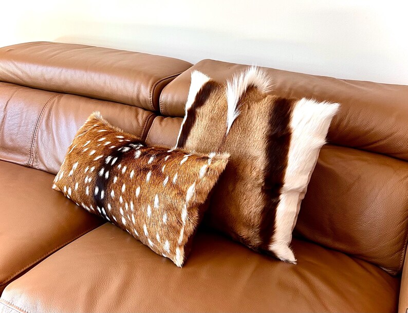 May include: Two decorative throw pillows with natural fur covers. One pillow features a brown and white spotted pattern, while the other has a brown, white, and black striped design. The pillows rest on a brown leather sofa.