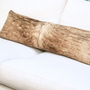 May include: A long, rectangular throw pillow with a brown and white cowhide pattern. The pillow is lying on a white couch.