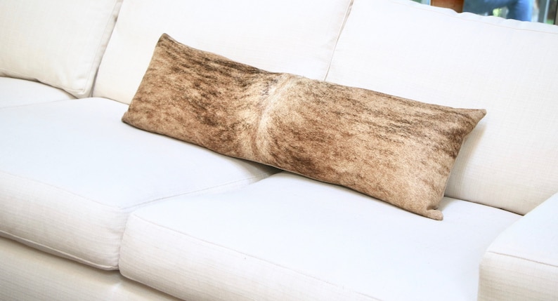 May include: A long, rectangular throw pillow with a brown and white cowhide pattern. The pillow is lying on a white couch.