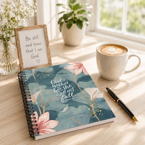 Puede incluir: Un cuaderno con espiral con un diseño floral y verde azulado. La portada presenta el texto "God is within her, she will not fall." También es visible una impresión enmarcada con el texto "Be still and know that I am God".