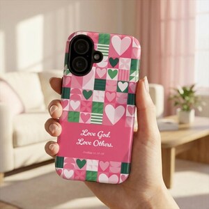 Może przedstawiać: Różowe etui na telefon z geometrycznym wzorem serc i kwadratów w odcieniach różu, zieleni i bieli. Etui zawiera tekst "Love God. Love Others." i werset biblijny Mateusza 22:37-39.
