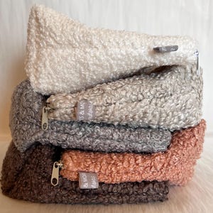 Könnte beinhalten: Ein Stapel von fünf Reißverschlusstaschen in verschiedenen neutralen Farben: Off-White, Hellgrau, Mittelgrau, Pfirsich und Dunkelbraun. Jede Tasche hat eine strukturierte, flauschige Außenseite und einen silbernen Reißverschluss. Die Taschen sind auf einer weißen, flauschigen Oberfläche gestapelt.