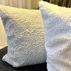 Könnte beinhalten: Zwei quadratische Kissen mit einem strukturierten, geschlungenen Stoff. Ein Kissen ist hellbeige, das andere weiß. Die Kissen liegen eng beieinander und zeigen die Textur des Stoffes.