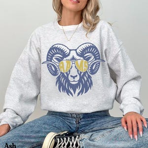 Op de afbeelding: Een lichtgrijs sweatshirt met ronde hals en een blauwe afbeelding van een ramshoofd met een gele zonnebril. De glazen van de zonnebril bevatten de tekst RAMS in een vetgedrukt lettertype. Het sweatshirt heeft lange mouwen en een boord.