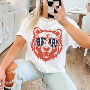 Pu&ograve; includere: Maglietta bianca con un orso rosso che indossa occhiali da sole blu con la scritta "BEARS" sulle lenti. L'orso indossa occhiali da sole.