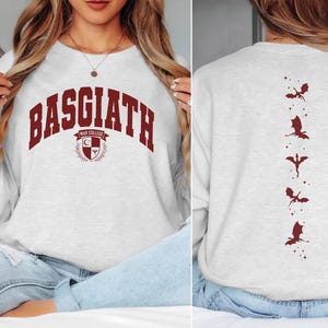 Puede incluir: Una sudadera gris con una impresión roja en la parte delantera que dice "BASGIATH WAR COLLEGE" y una impresión roja de cinco dragones en la parte posterior. La sudadera se muestra en una modelo que lleva pantalones vaqueros azules.
