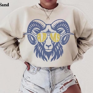 May include: A sand-colored crewneck sweatshirt featuring a large graphic of a ram head wearing yellow-tinted sunglasses. The ram is rendered in a blue line-art style, and the word RAMS is printed in yellow across the lenses of the sunglasses.
