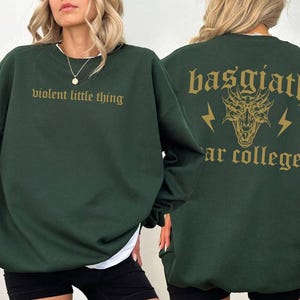 Pode incluir: Uma camisa de moletom verde escura com o texto "violent little thing" na frente e "basgiath" com um gr&aacute;fico de drag&atilde;o e o texto "ar college" nas costas.