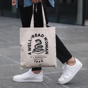 Pode incluir: Uma bolsa tote de lona branca com texto preto que diz "A well-read woman is a dangerous creature 1920". A bolsa apresenta uma ilustração em preto e branco de uma cobra enrolada em torno de um livro.