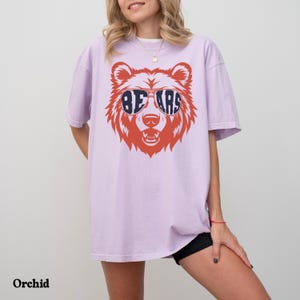 Pu&ograve; includere: Una maglietta color lilla chiaro con una grafica di un orso bruno che indossa occhiali da sole con la scritta "BEARS" in lettere blu.
