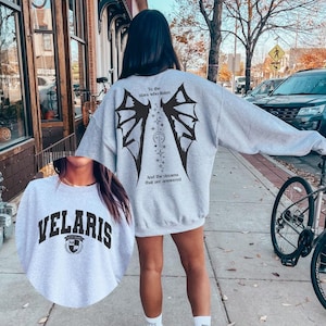 May include: A light gray sweatshirt with black graphic wings and text that reads "To the stars who listen" and "And the dreams that are answered." A smaller image shows a gray sweatshirt with the word "VELARIS" in black.