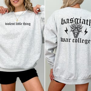 Pode incluir: Camisola de moletom cinza com o texto "violent little thing" na frente e "basgiath war college" com um gr&aacute;fico de drag&atilde;o nas costas.