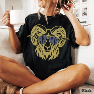 May include: A black t-shirt featuring a gold graphic of a ram head wearing blue aviator sunglasses. The lenses of the sunglasses contain the text RAMS in a bold, dark blue font. The ram has large, curved horns and a detailed, stylized face.
