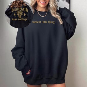 Pode incluir: Camisola de moletom preta oversized com o texto "violent little thing" em letras douradas. A camisola tem gola redonda e mangas compridas.