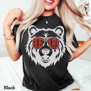 Pu&ograve; includere: Maglietta nera con un'immagine di un orso bianco che indossa occhiali da sole rossi con la scritta "BEARS".