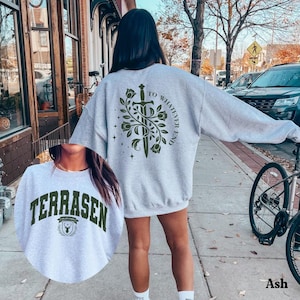 Puede incluir: Una sudadera gris claro con la palabra "TERRASEN" en letras de bloque verde oscuro. La sudadera también presenta un diseño en la espalda con una espada, flores y el texto "TO WHATEVER END" en verde oscuro.