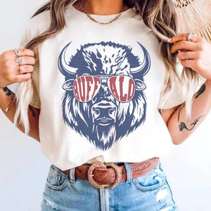May include: An ivory short-sleeve t-shirt featuring a graphic print of a blue buffalo wearing red-rimmed sunglasses. The word BUFFALO is printed in white letters across the lenses of the sunglasses. The shirt has a crew neckline and a relaxed fit.