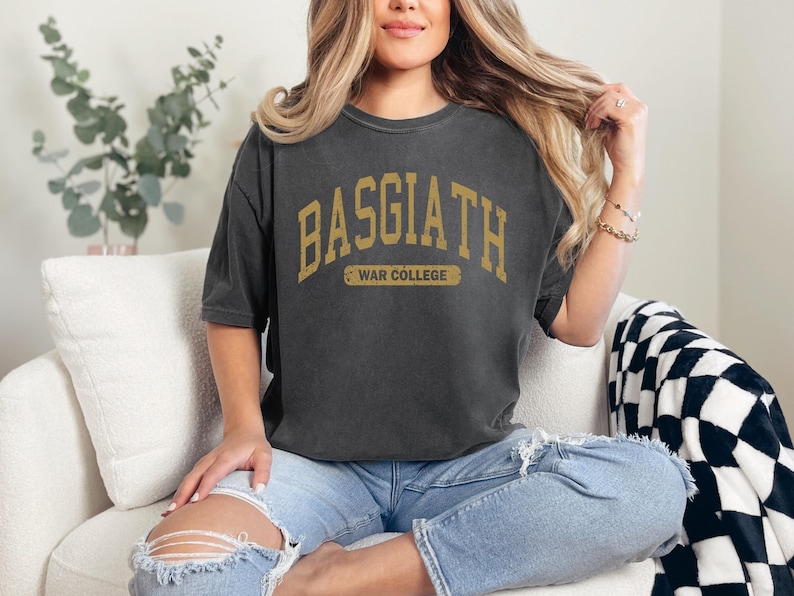 Op de afbeelding: Een donkergrijs T-shirt met een vintage-stijl grafische print met de tekst "Basgiath War College" in gouden letters.