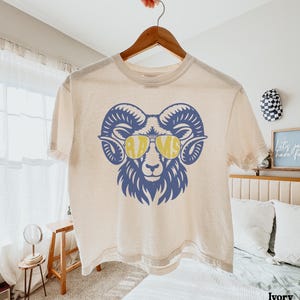 May include: An ivory short-sleeved t-shirt featuring a blue graphic of a ram head wearing yellow aviator sunglasses. The lenses of the sunglasses contain the text RAMS in a bold, stylised font. The shirt has a crew neckline and a relaxed fit.