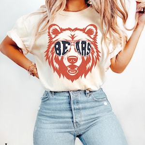 Pu&ograve; includere: Una maglietta bianca con un orso rosso che indossa occhiali da sole che dicono "BEARS" in lettere blu. La maglietta &egrave; indossata da una persona con capelli lunghi e biondi e jeans blu.