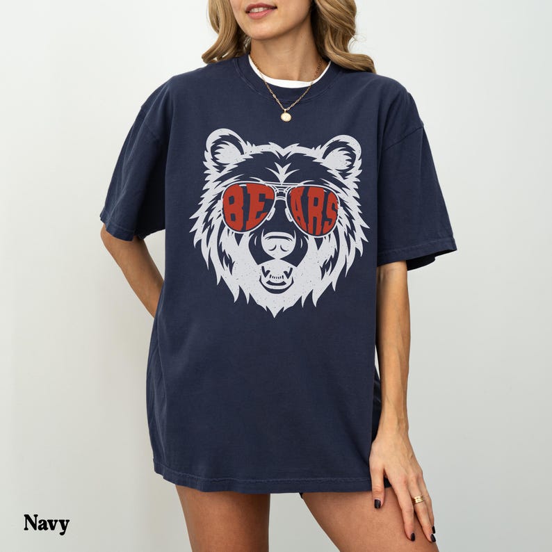 Pu&ograve; includere: Una t-shirt blu navy con un grafico di un orso bianco che indossa occhiali da sole che dicono "BEARS" in lettere rosse.