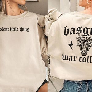 Pode incluir: Duas sweatshirts bege com designs gr&aacute;ficos diferentes. Uma sweatshirt diz "violent little thing" e a outra diz "basgiath war college" com um gr&aacute;fico de drag&atilde;o.
