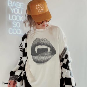 Könnte beinhalten: Ein elfenbeinfarbenes T-Shirt mit einem schwarzen Grafikdruck von Lippen mit Vampirzähnen. Das Outfit umfasst eine senfgelbe Trucker-Cap mit dem Text MAD LOVE FOR YOU und eine schwarz-weiß karierte Strickjacke mit langen Ärmeln.
