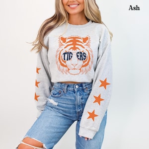 May include: An ash grey long-sleeve sweatshirt featuring a large orange tiger graphic on the chest. The tiger wears sunglasses with the text TIGERS printed on the lenses. Each sleeve is decorated with a vertical row of three orange stars.