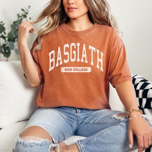 Op de afbeelding: Een roestkleurig T-shirt met de tekst "BASGIATH WAR COLLEGE" in wit gedrukt. Het shirt is gemaakt van een zacht, comfortabel materiaal en heeft een ontspannen pasvorm.