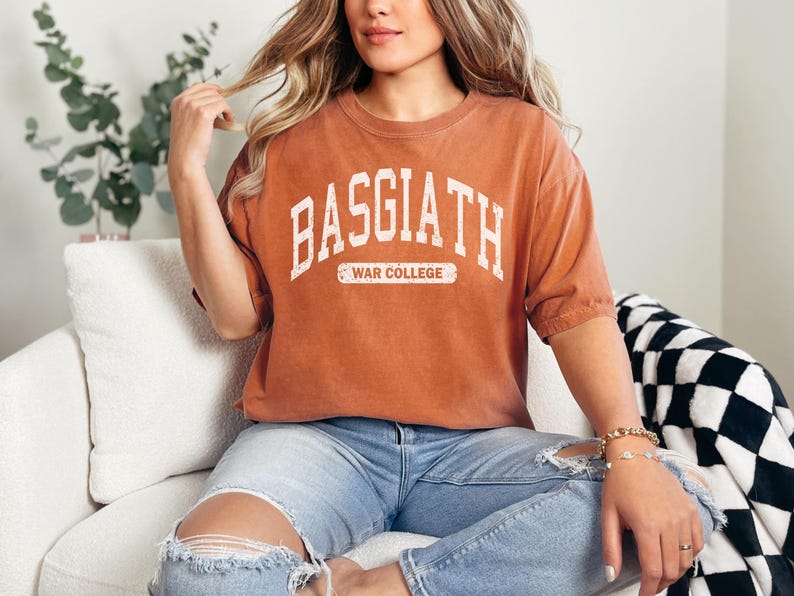 Op de afbeelding: Een roestkleurig T-shirt met de tekst "BASGIATH WAR COLLEGE" in wit gedrukt. Het shirt is gemaakt van een zacht, comfortabel materiaal en heeft een ontspannen pasvorm.