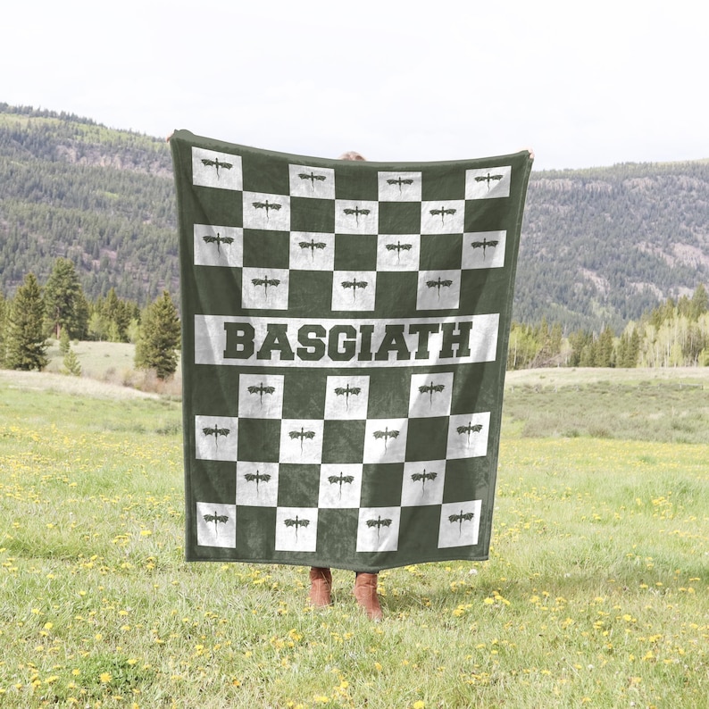 K&ouml;nnte beinhalten: Eine gr&uuml;ne Fleece-Decke mit einem karierten Muster aus wei&szlig;en und gr&uuml;nen Quadraten. Jedes Quadrat zeigt ein schwarzes Libellen-Design. Die Decke hat das Wort "BASGIATH" in wei&szlig;en Buchstaben in der Mitte aufgedruckt.