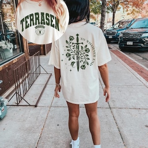 May include: Ivory-colored t-shirt with the word "TERRASEN" in dark green block letters. The back of the shirt features a sword design with the text "TO WHATEVER END" in dark green. The shirt is worn outdoors.