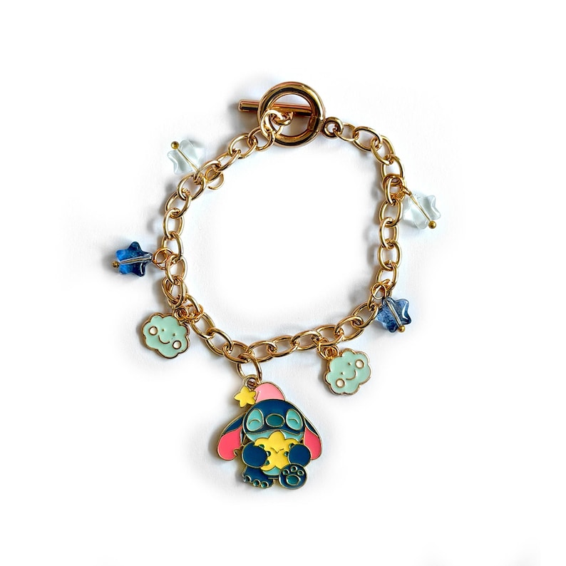 May include: Gold-toned chain bracelet with a toggle clasp. The bracelet features charms of a blue cartoon character, blue stars, and light blue clouds. The cartoon character is holding a yellow star. The bracelet is on a white background.