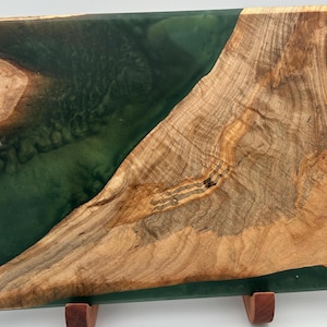 May include: A rectangular wooden serving tray with a live edge design. The tray features a section of deep green epoxy resin and is accented with gold-colored handles. The wood has a natural, light brown color.