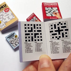 K&ouml;nnte beinhalten: Miniatur-Puzzle-B&uuml;cher, darunter ein Sudoku und mehrere Kreuzwortr&auml;tsel. Das Sudoku-Buch ist beige mit schwarzem Text und einem roten Kreis. Die Kreuzwortr&auml;tselb&uuml;cher sind rot und blau mit schwarz-wei&szlig;en Kreuzwortr&auml;tseln.