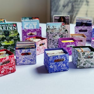 May include: A collection of miniature storage boxes in various colors, including red, blue, green, and purple. Each box features a leaf pattern and holds small stacks of paper or magazines. Some boxes have magazine holders.