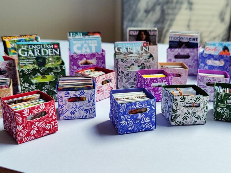 May include: A collection of miniature storage boxes in various colors, including red, blue, green, and purple. Each box features a leaf pattern and holds small stacks of paper or magazines. Some boxes have magazine holders.