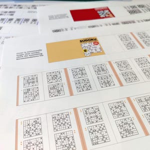 K&ouml;nnte beinhalten: Eine Sammlung von gedruckten Sudoku-R&auml;tseln und anderen Dokumenten. Die Sudoku-R&auml;tsel sind in einem Rasterformat mit Zahlen angeordnet. Das Bild enth&auml;lt auch einen gelben Umschlag mit dem Aufdruck "Sudoku" sowie anderen Text und QR-Codes.