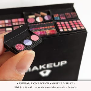May include: A miniature black makeup display stand featuring various small cosmetic palettes and lipstick sets. A single open black compact with three circular colored pans is held in the foreground. The base of the stand displays the word MAKEUP.