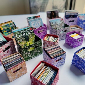 May include: Miniature decorative storage boxes in various colors and patterns, filled with tiny magazines. The boxes feature floral and leaf designs in shades of green, purple, red, and blue. Some magazines are visible, with titles like "The English Garden" and "What Your Dog Is Trying to Tell You."