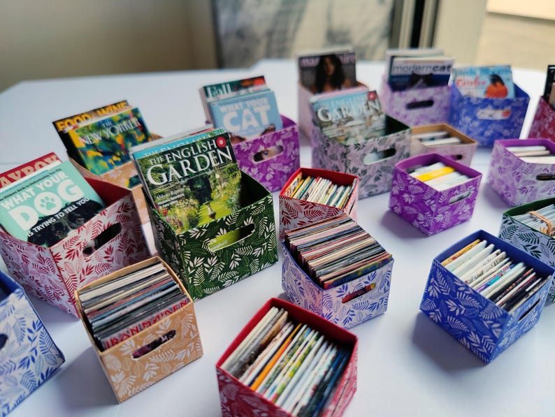 May include: Miniature decorative storage boxes in various colors and patterns, filled with tiny magazines. The boxes feature floral and leaf designs in shades of green, purple, red, and blue. Some magazines are visible, with titles like "The English Garden" and "What Your Dog Is Trying to Tell You."