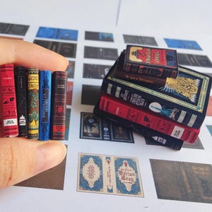 May include: Miniature books in various colors, including red, black, blue, and brown. The books are stacked and held by a hand. Titles include "Dracula" and "Classic Stories". The books are on a white surface with other printed designs.