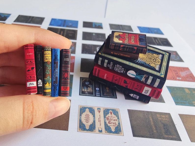 May include: Miniature books in various colors, including red, black, blue, and brown. The books are stacked and held by a hand. Titles include "Dracula" and "Classic Stories". The books are on a white surface with other printed designs.