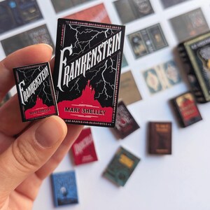 May include: Miniature book replicas of "Frankenstein" by Mary Shelley. The books have black covers with white lettering and red accents. Lightning bolts and a castle silhouette are featured on the cover. Other miniature books are scattered in the background.