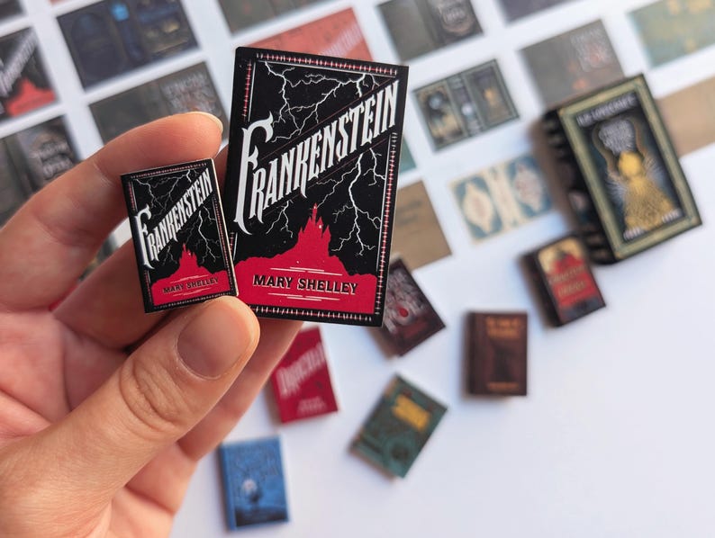 May include: Miniature book replicas of "Frankenstein" by Mary Shelley. The books have black covers with white lettering and red accents. Lightning bolts and a castle silhouette are featured on the cover. Other miniature books are scattered in the background.