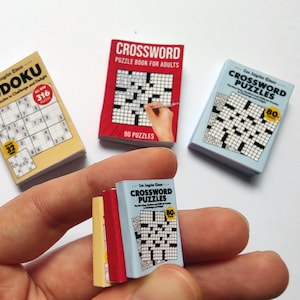 K&ouml;nnte beinhalten: Eine Hand h&auml;lt Miniatur-Puzzleb&uuml;cher, darunter Sudoku- und Kreuzwortr&auml;tsel. Die B&uuml;cher haben farbenfrohe Einb&auml;nde mit Texten wie "Los Angeles Times" und "Crossword Puzzles". Das Sudoku-Buch enth&auml;lt 316 R&auml;tsel.
